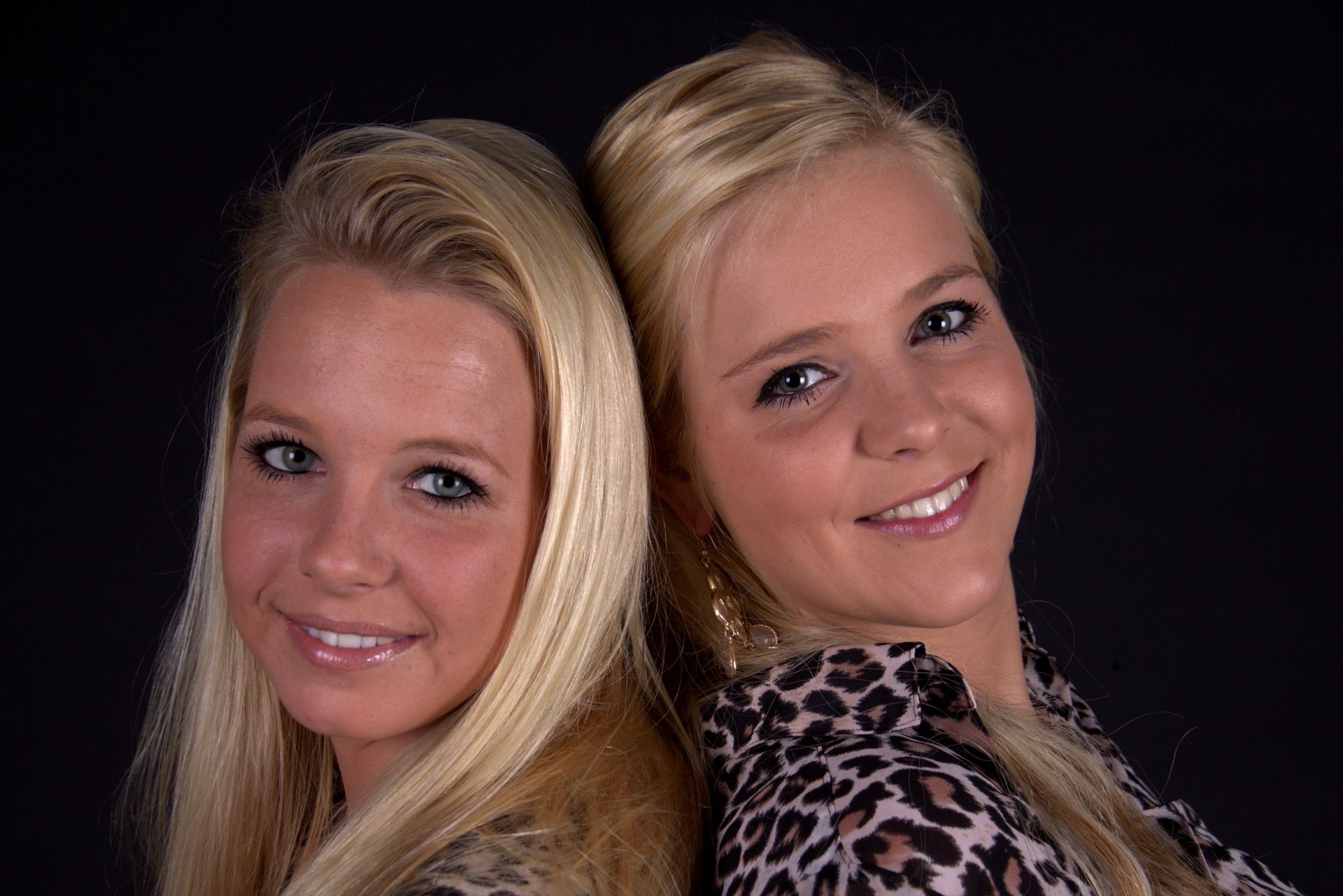 Twee lachende blonde vrouwen poseren met hun ruggen tegen een donkere achtergrond.