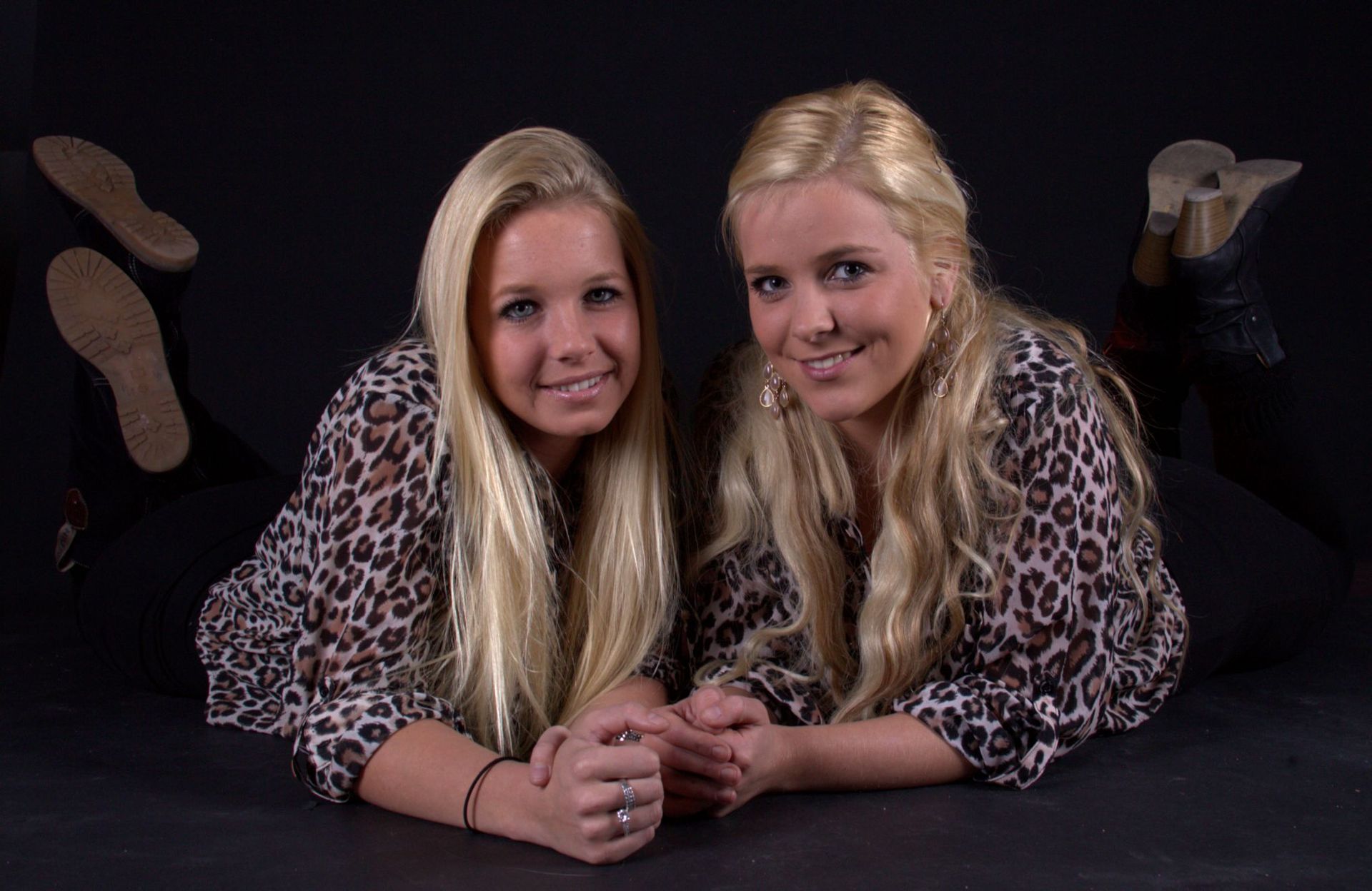 Twee vrouwen liggen lachend op een donkere vloer, gekleed in topjes met luipaardprint en zwarte broeken.