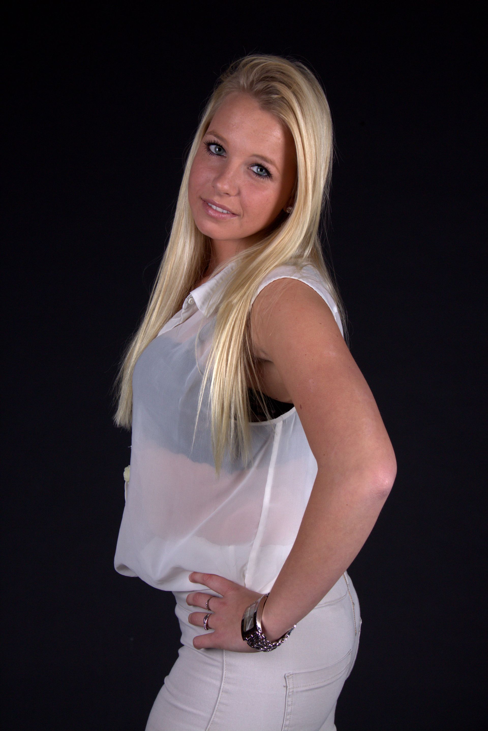 Blonde vrouw poseert tegen een donkere achtergrond, gekleed in een witte top en een zwarte broek.