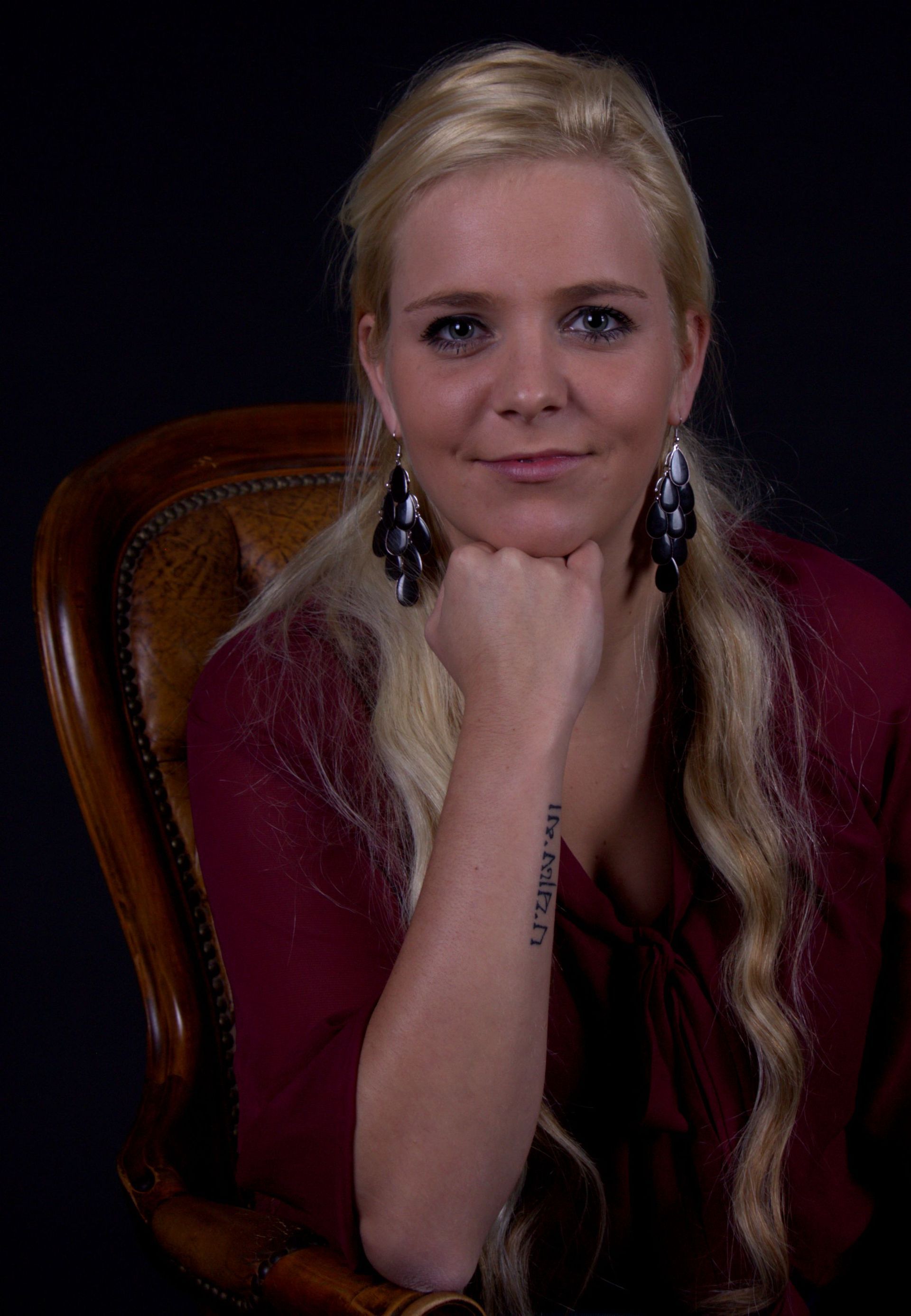 Portret van een vrouw met lang blond haar, die haar kin op haar hand laat rusten tegen een donkere achtergrond.