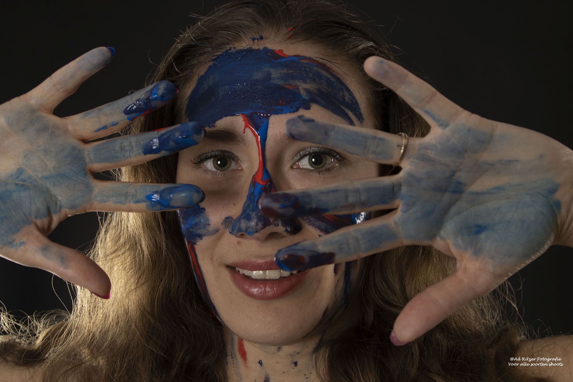 Vrouw met blauwgeverfde handen die haar gezicht omlijsten, blauwe en rode verf rond haar ogen en neus, glimlachend.
