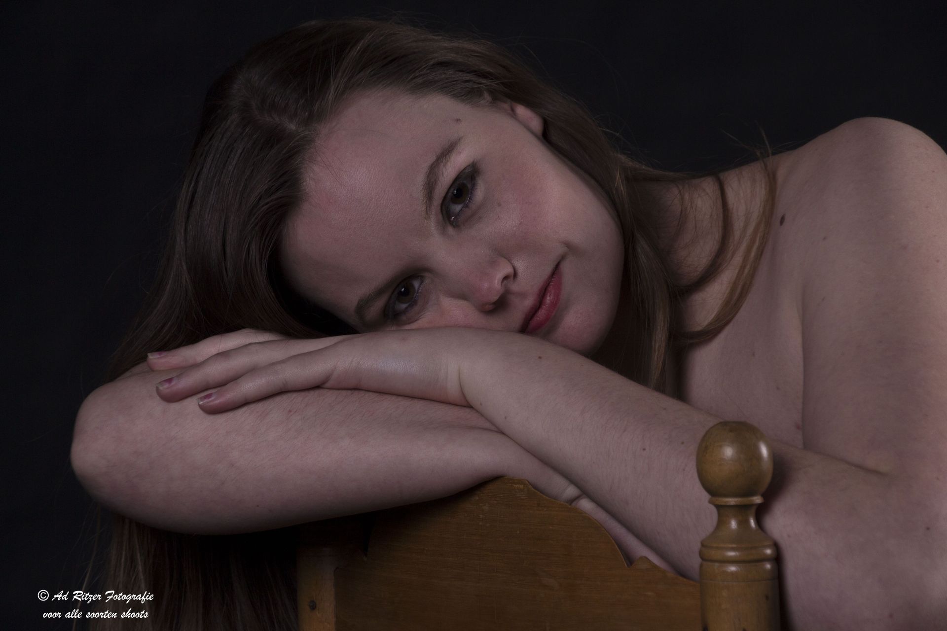 Een vrouw laat haar hoofd rusten op haar gevouwen armen, leunend tegen een houten stoel, en kijkt in het zachte licht naar de camera.