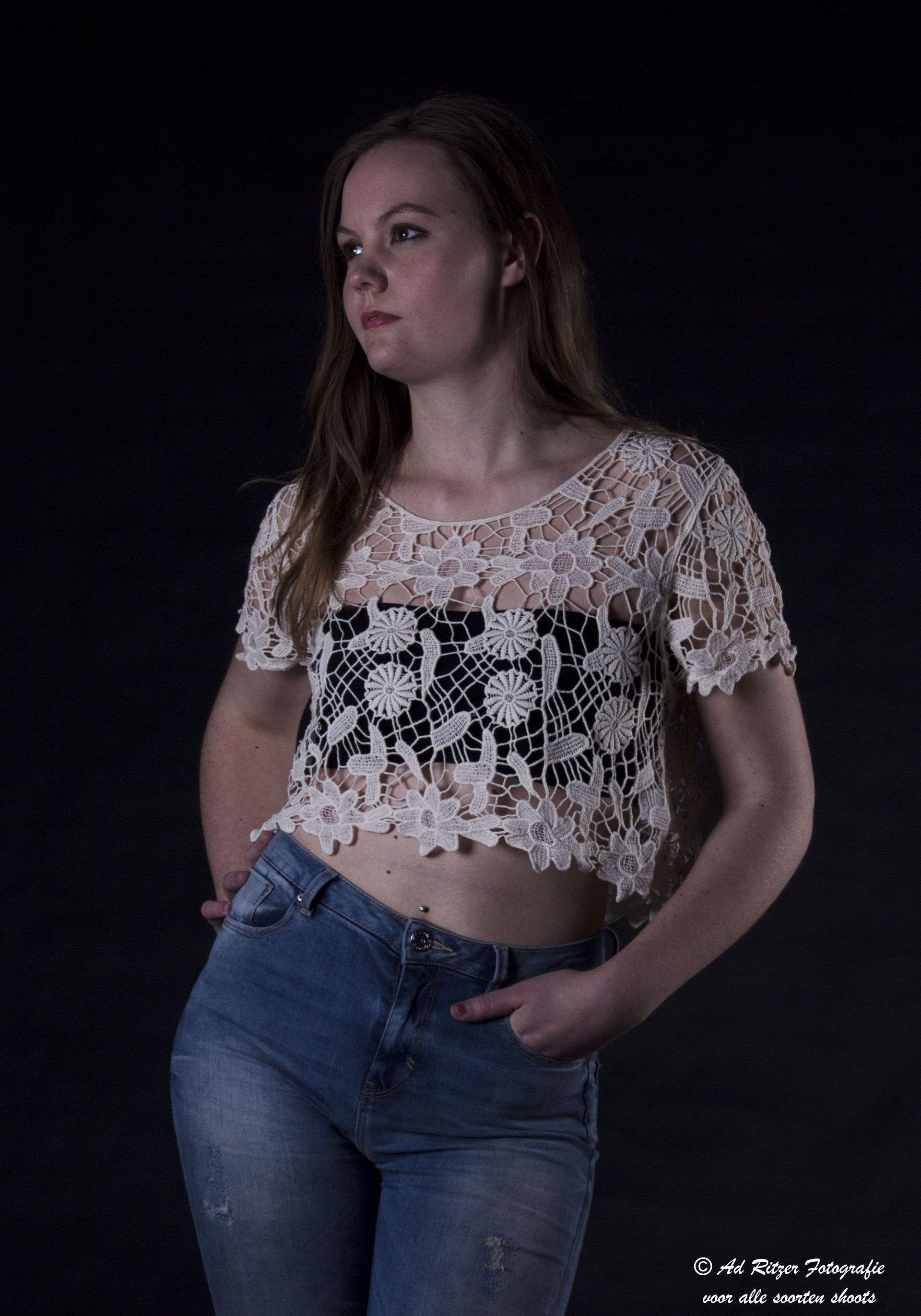 Vrouw in een kanten crop top en jeans poseert tegen een donkere achtergrond, handen in de zakken.