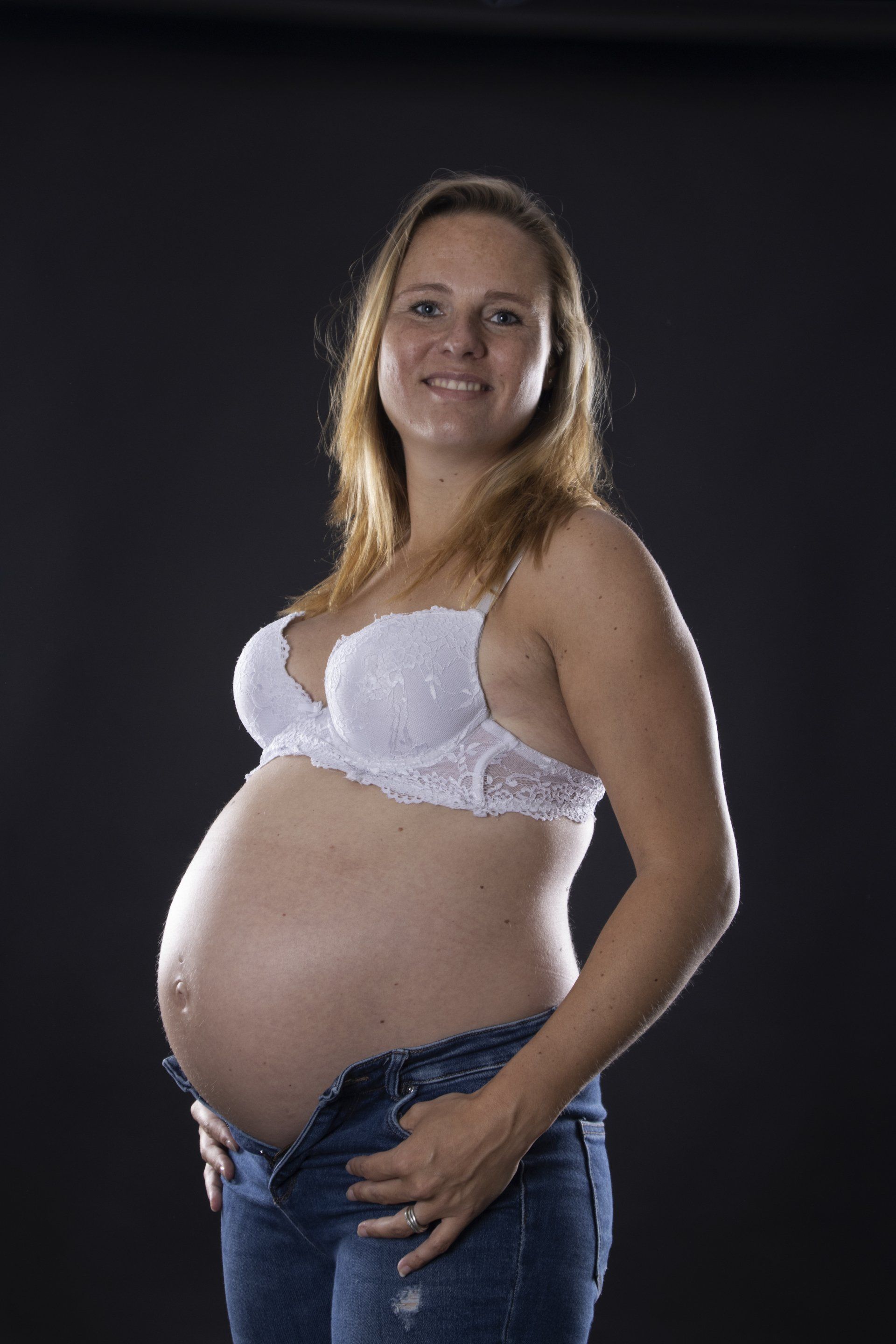Een zwangere vrouw staat tegen een donkere achtergrond, gekleed in een witte bh en een blauwe spijkerbroek.