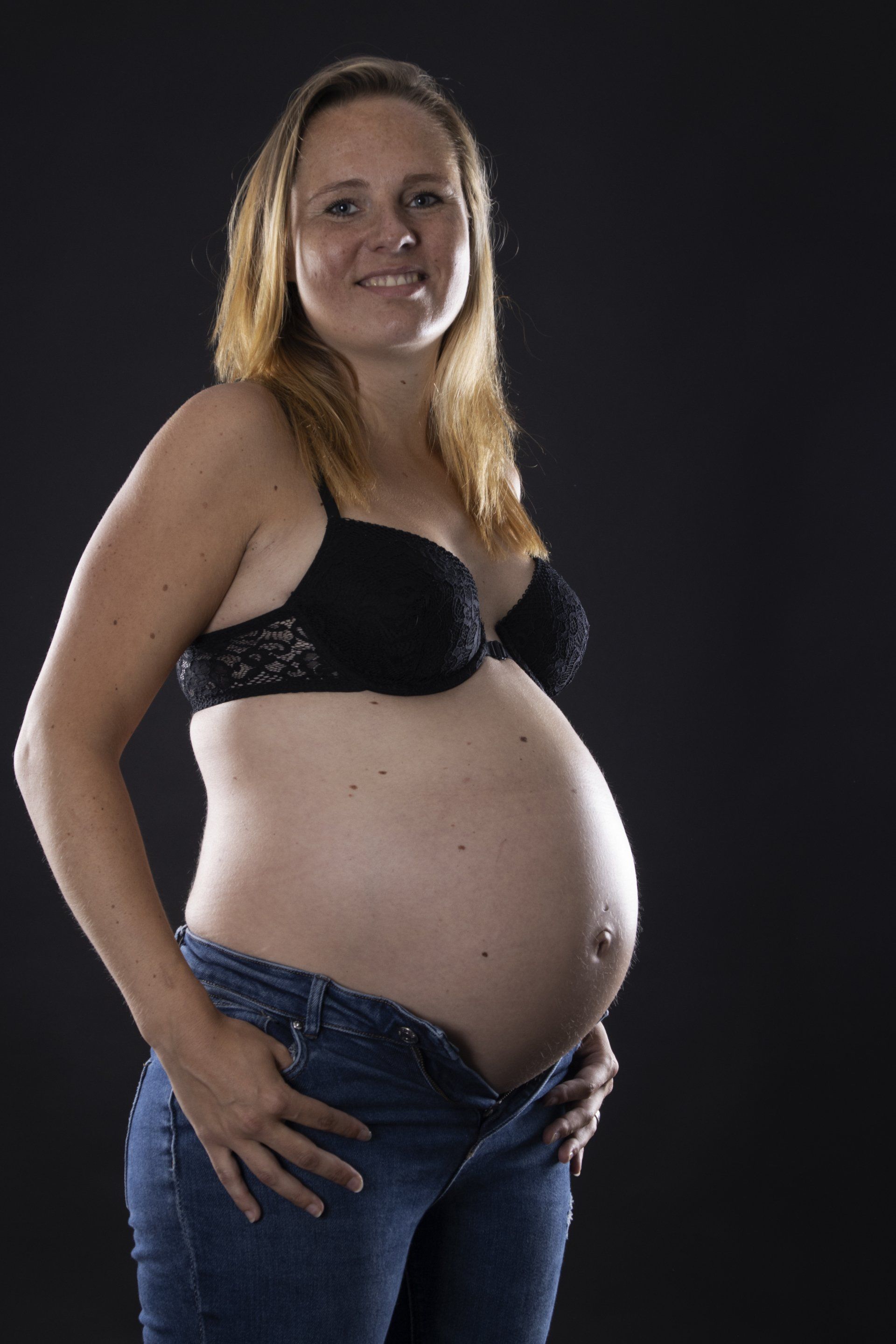 Zwangere vrouw poseert in een zwarte bh en jeans tegen een donkere achtergrond.