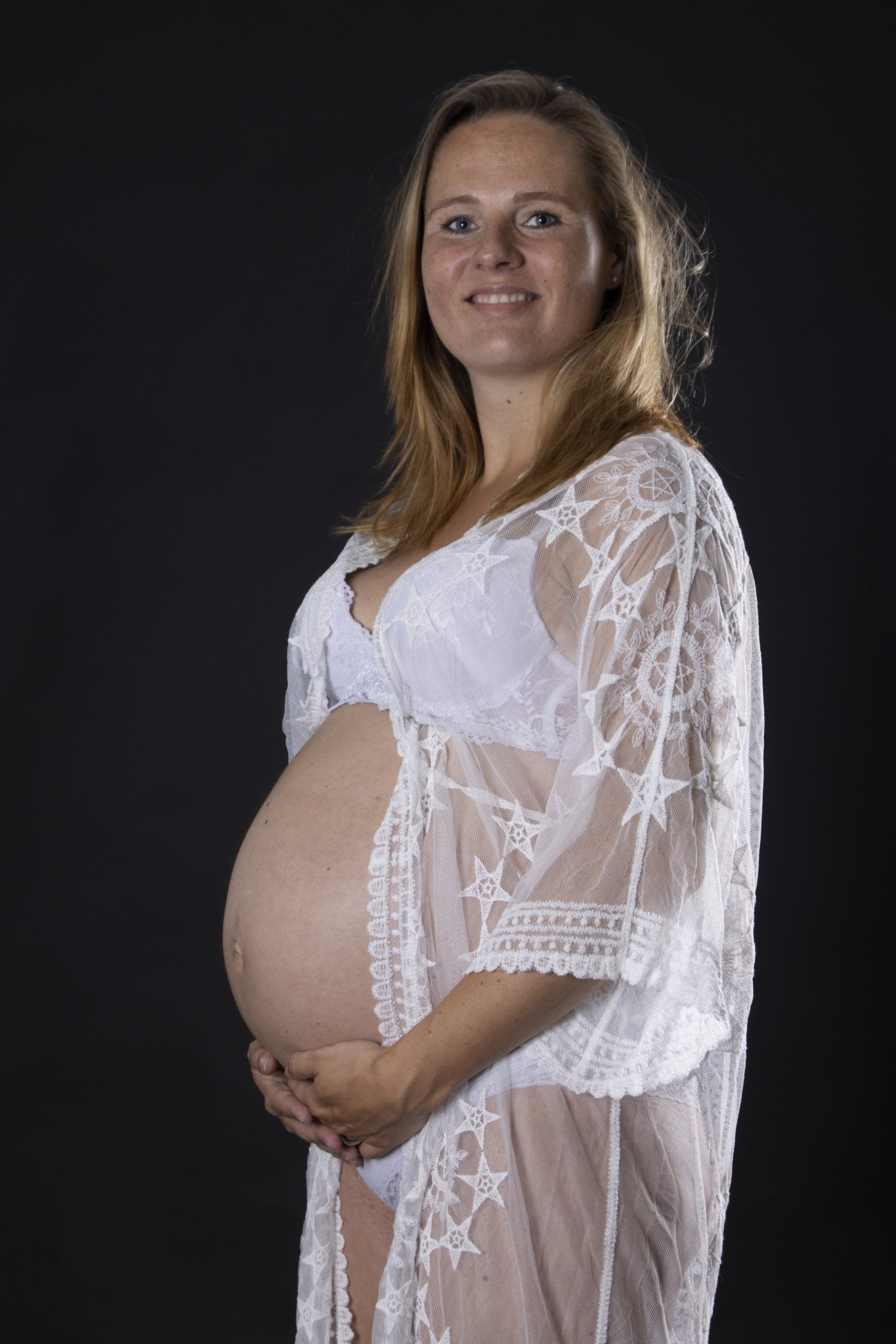 Een zwangere vrouw in een witte kanten jurk staat in profiel tegen een donkere achtergrond, met haar handen op haar buik.