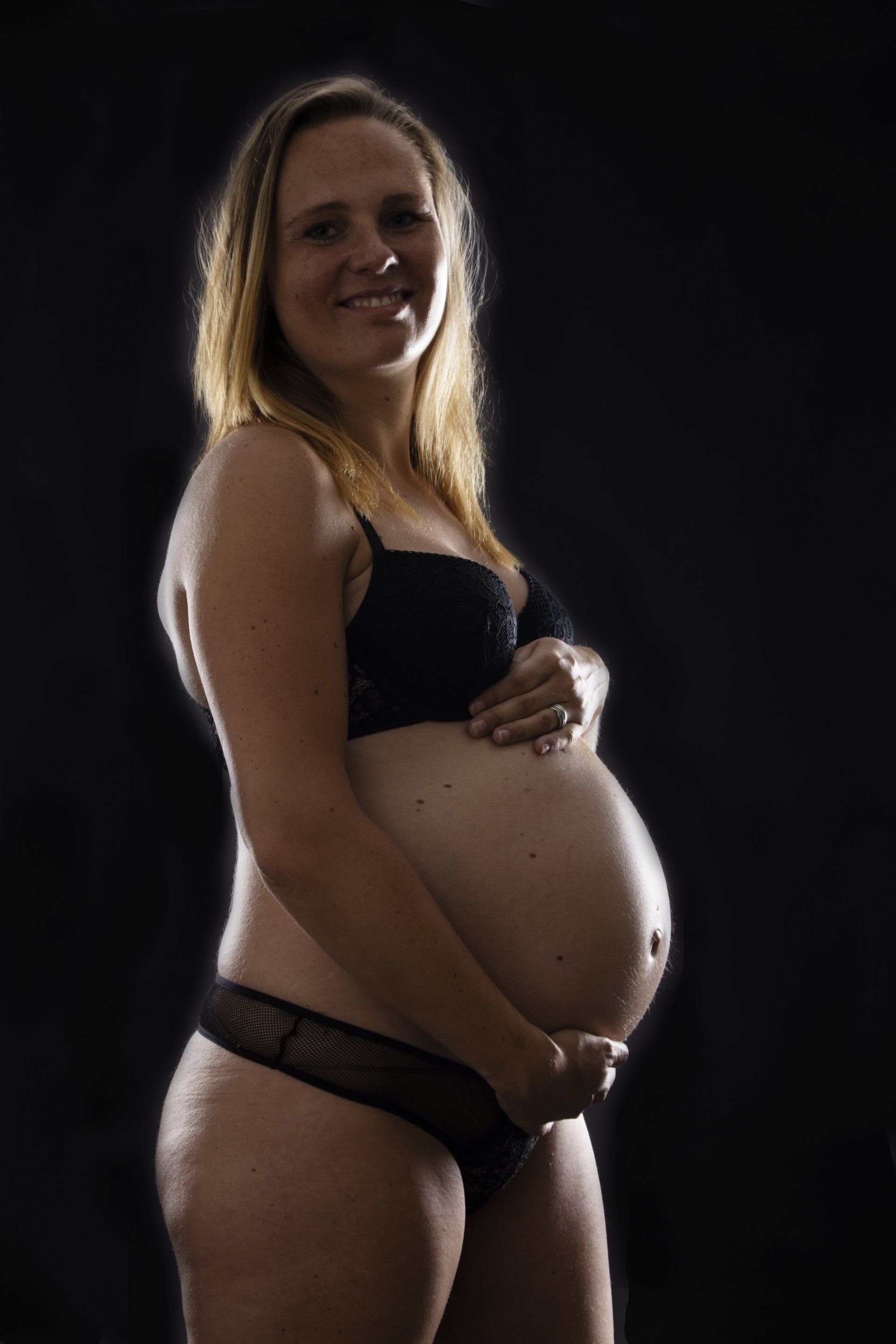 Een zwangere vrouw in zwarte lingerie poseert tegen een donkere achtergrond, lachend en haar buik vasthoudend.
