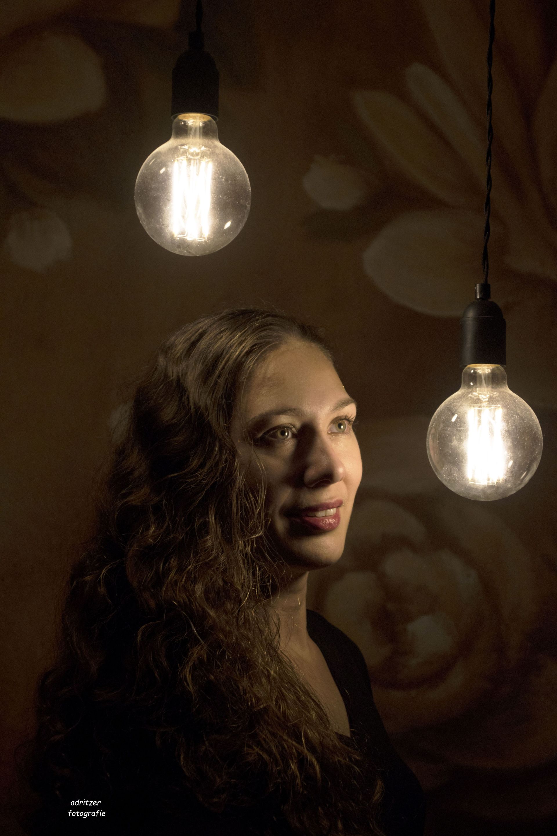 Vrouw in een donkere studio met twee gloeiende hanglampen, kijkt opzij.