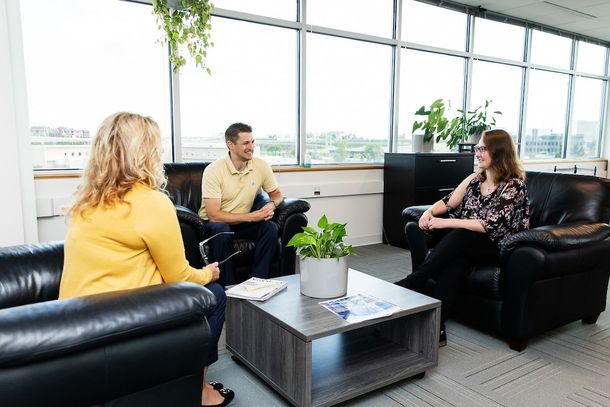 Three people in a bright office lounge having a conversation on black leather chairs.
