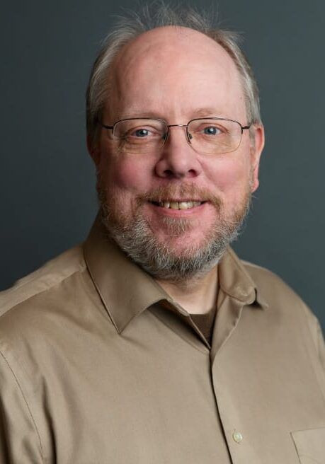 Studio portrait of a smiling man with glasses, wearing a tan shirt on a gray background