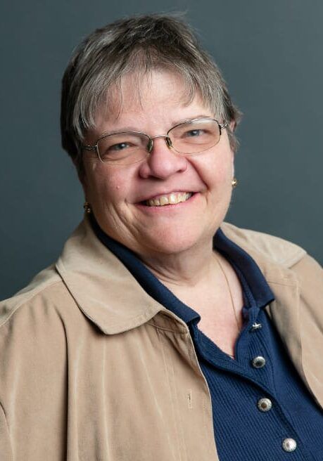 Smiling person in glasses wearing a beige jacket and navy shirt against a gray background