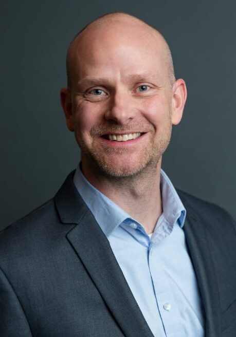 Smiling bald man in a blue shirt and dark blazer against a gray background