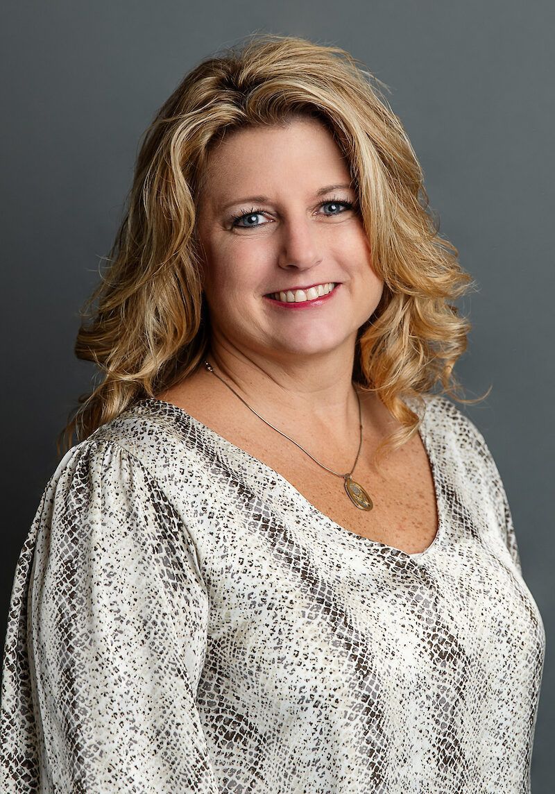 Smiling woman with shoulder-length blonde hair in a patterned blouse against a gray backdrop