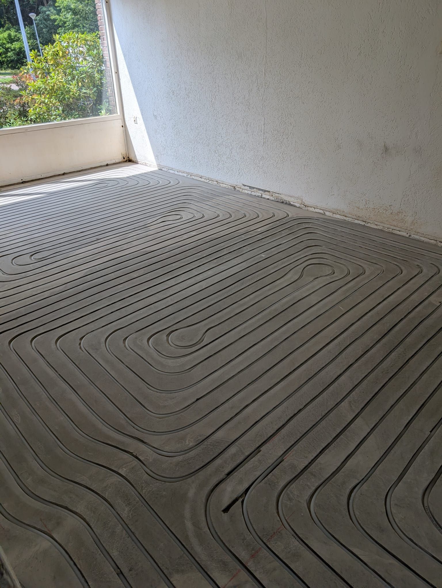 Betonnen vloer met golvende troffelgroeven naast een witte muur en een deuropening die uitkomt op groen.