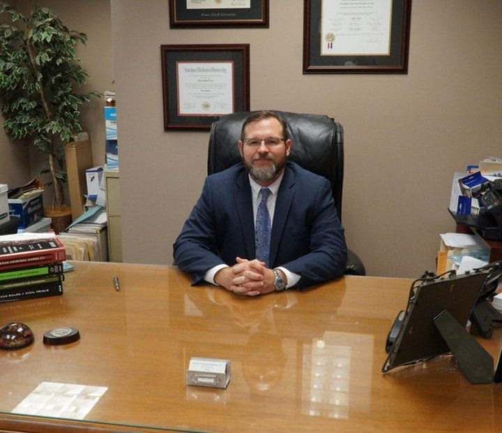 A man in a suit and tie sits at a desk with his hands folded