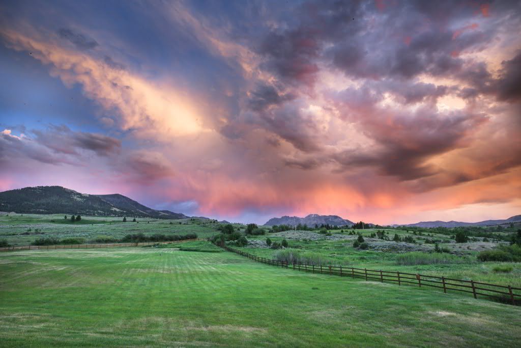 A sunset over a grassy field with a fence in the foreground and mountains in the background.