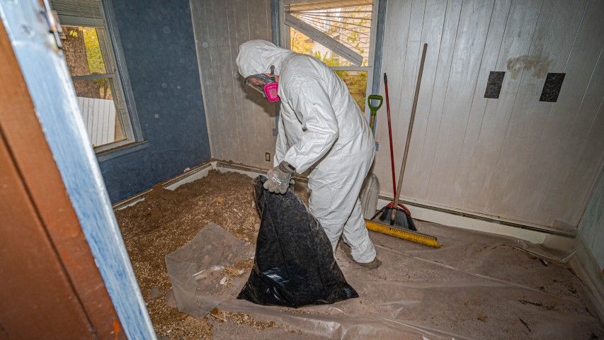 Person in white coveralls vacuuming debris in a dusty room with exposed walls and tools.