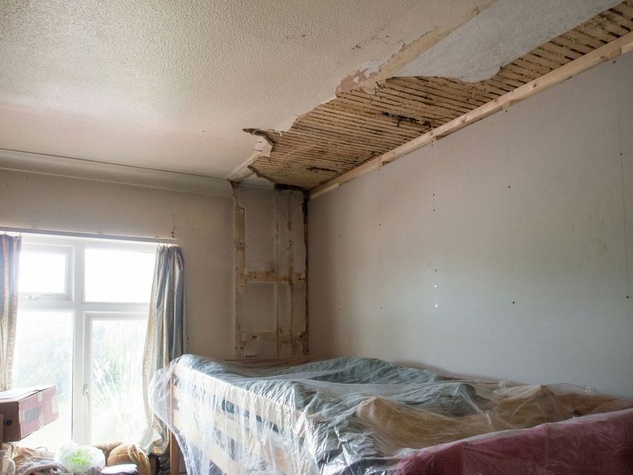 Damaged bedroom ceiling with exposed plaster and a bed covered in plastic near a window