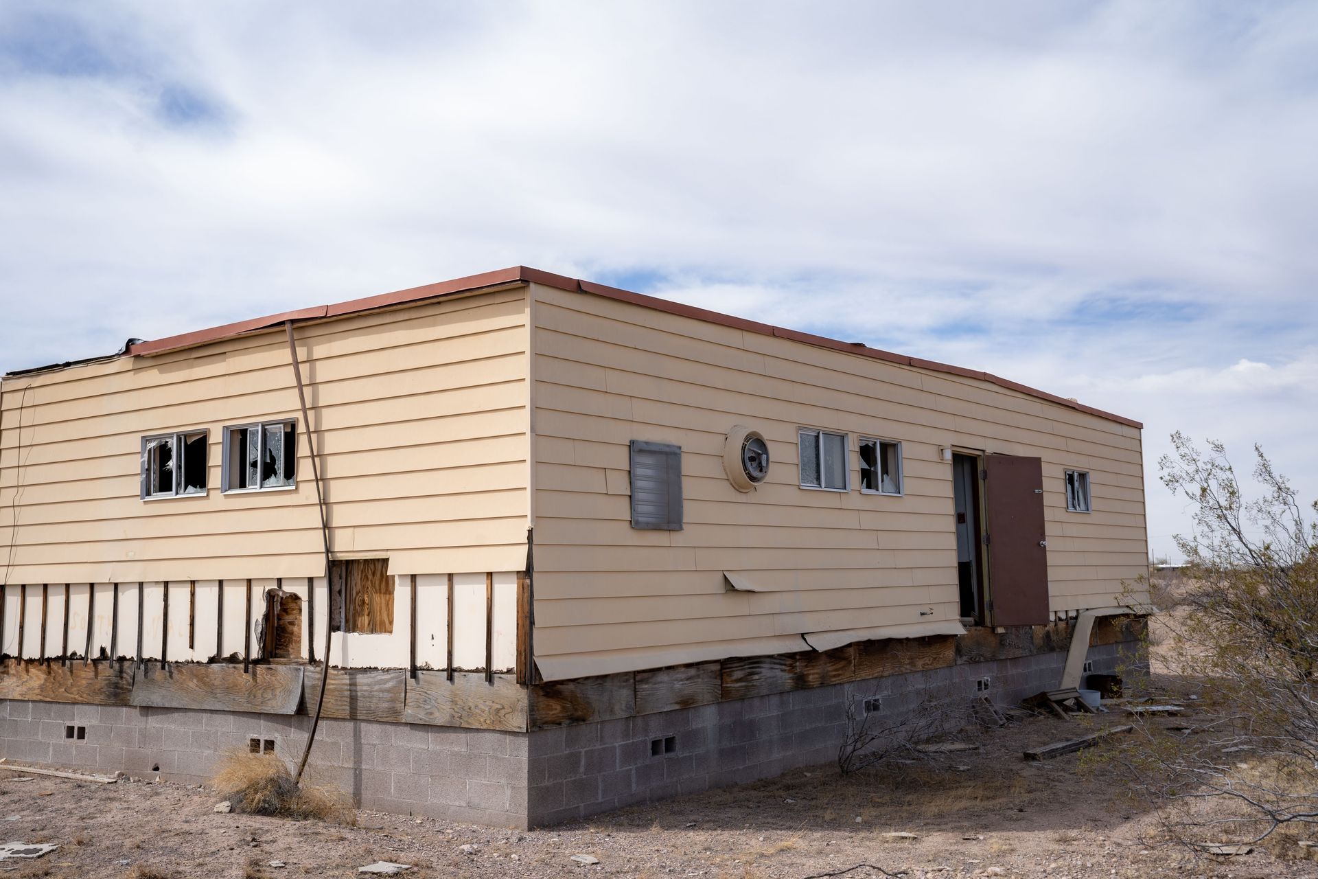 Weathered beige trailer house on a bare, rocky lot under a cloudy sky
