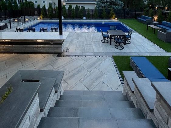 Outdoor patio with pool. Steps lead to a grey stone paver patio, dining area, and pool with blue water. Green turf grass surrounds.