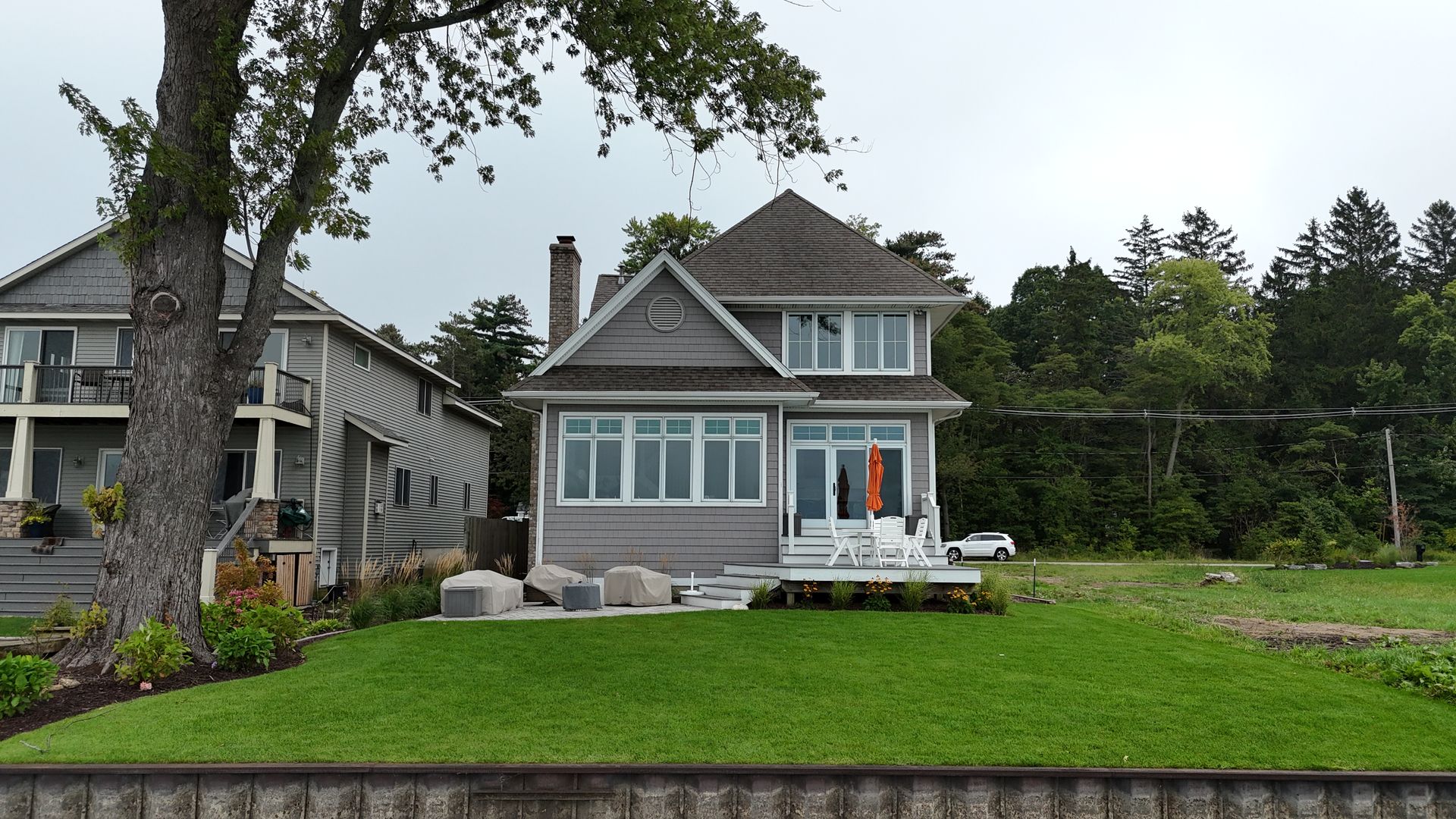 Gray cottage with shingle roof on a grassy waterfront, white deck, trees in background.