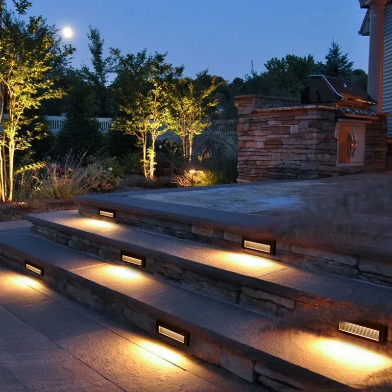 Stone steps illuminated by warm, rectangular lights at night, leading to an outdoor kitchen and trees.