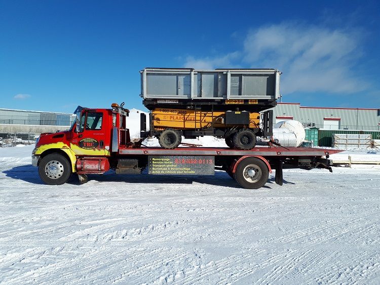 Camion de dépannage rouge et blanc avec une remorque jaune chargée sur son plateau, dans un décor extérieur enneigé.