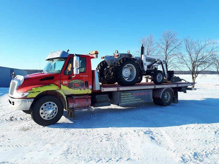 Une dépanneuse rouge transporte un tracteur argenté par une journée de neige.