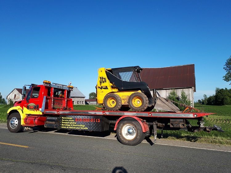 Camion de dépannage rouge transportant une chargeuse compacte JCB jaune et noire sur une remorque à plateau.