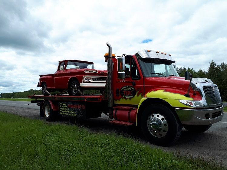 Une dépanneuse rouge transporte une camionnette classique rouge en bord de route, sous un ciel nuageux.