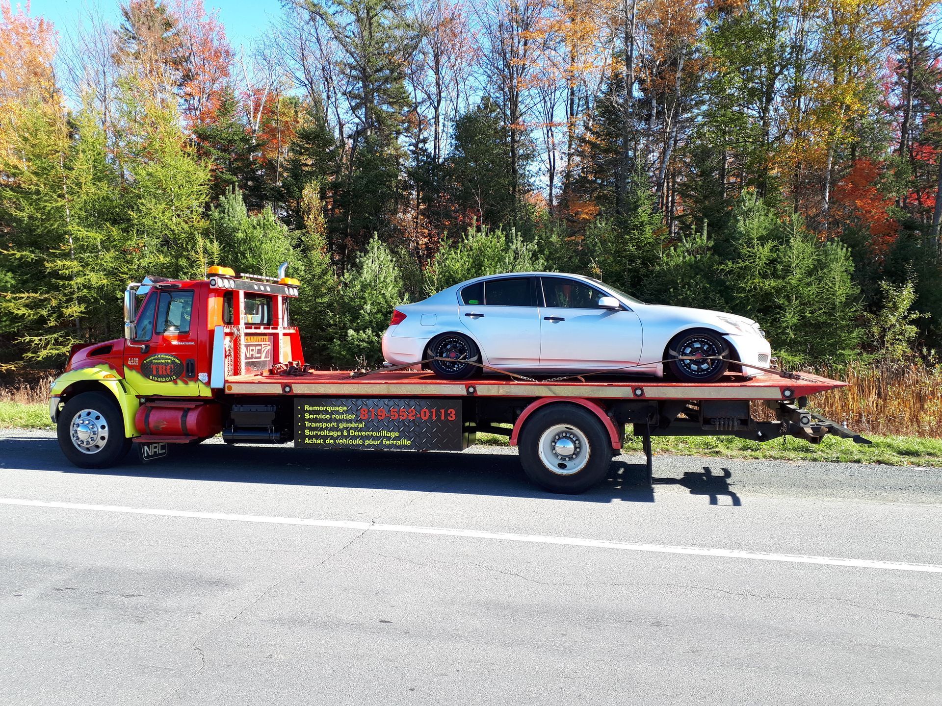 Une dépanneuse avec une berline argentée sur son plateau, garée en bord de route avec des feuillages d'automne.