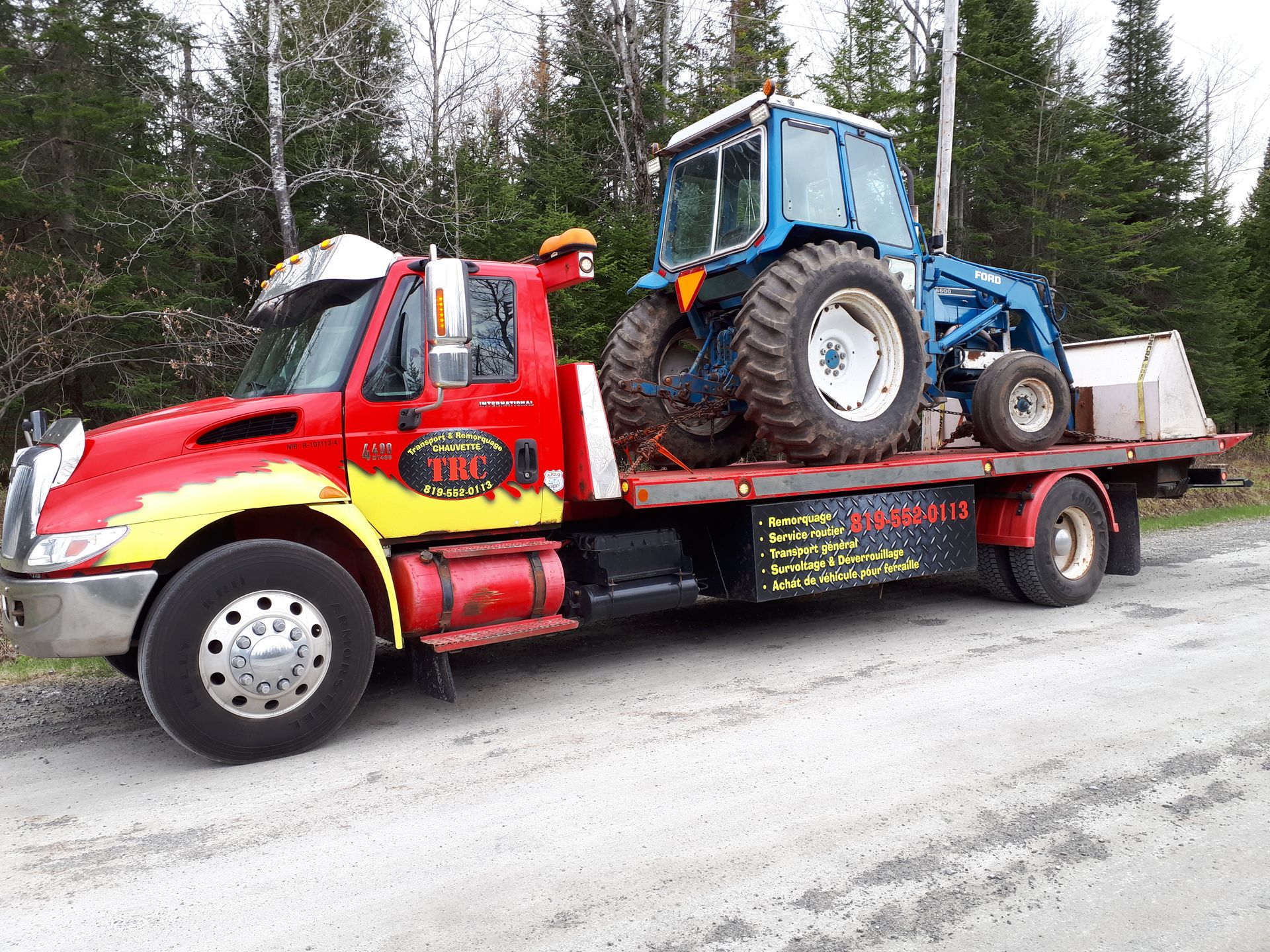 Un camion de dépannage rouge et jaune transporte un tracteur bleu sur une remorque à plateau, dans une zone boisée.