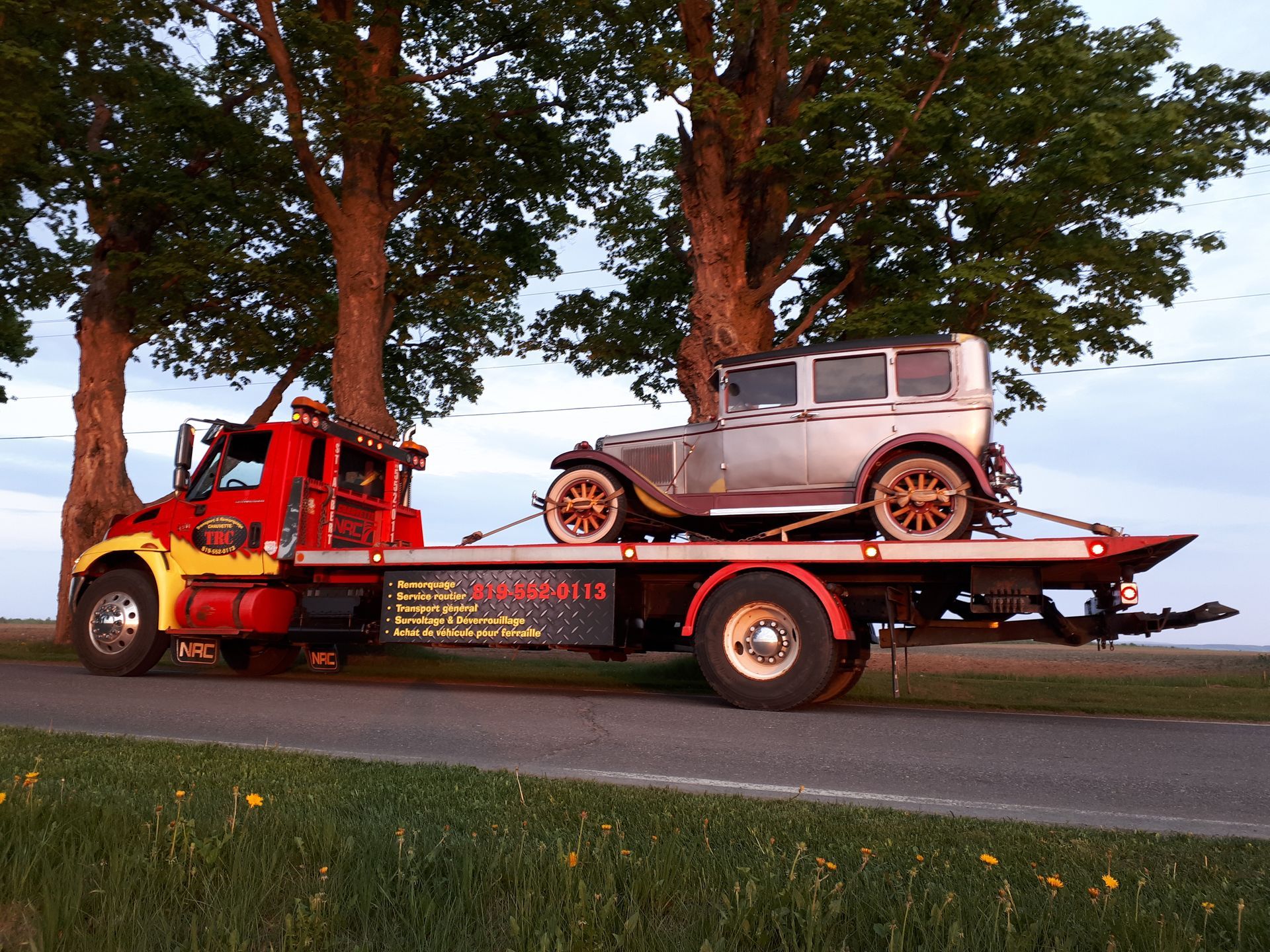 Dépanneuse transportant une voiture argentée de collection sur une remorque à plateau, en bord de route.