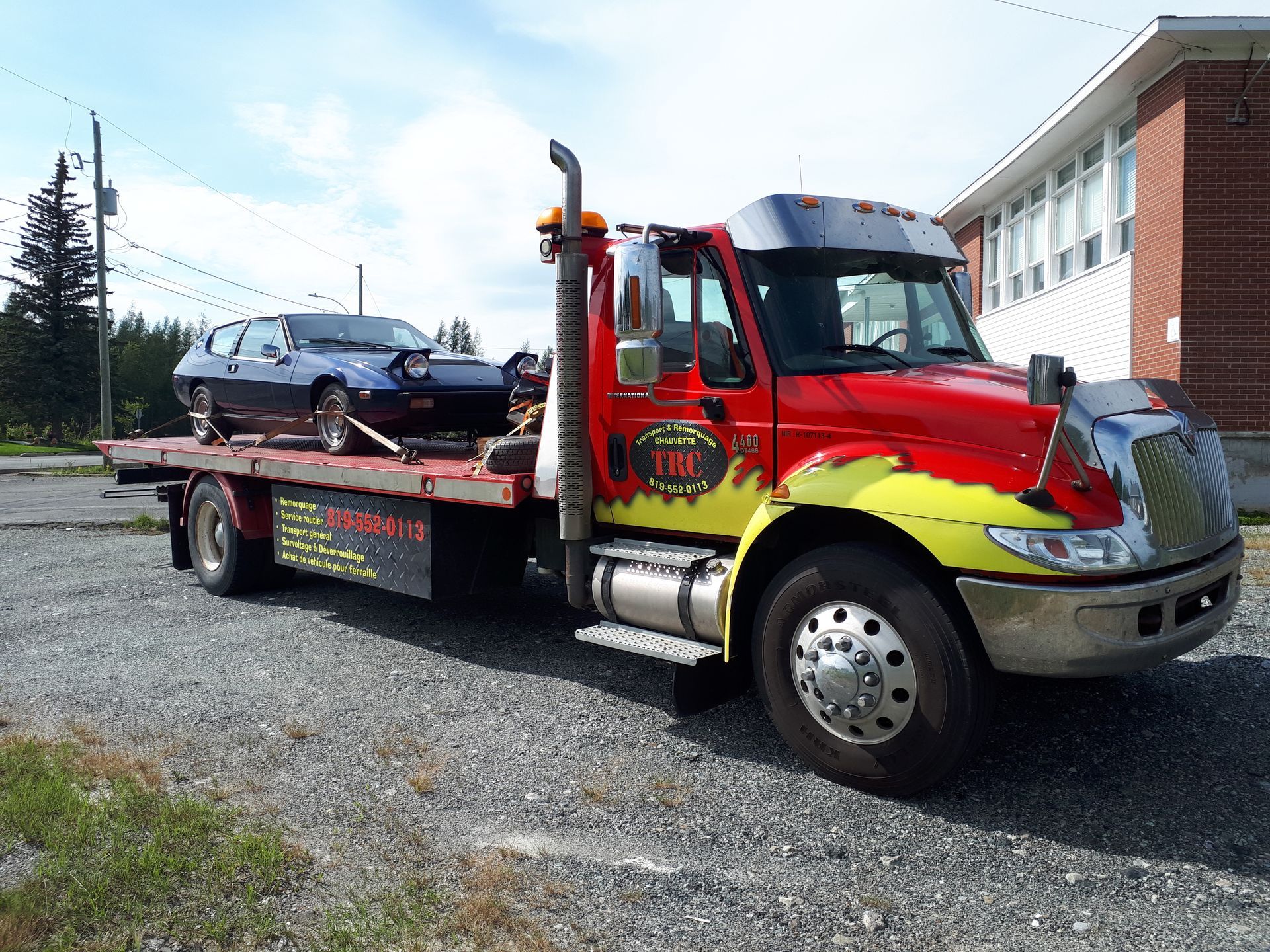 Une dépanneuse avec une voiture bleue sur son plateau, stationnée sur du gravier. Le camion est rouge et jaune.