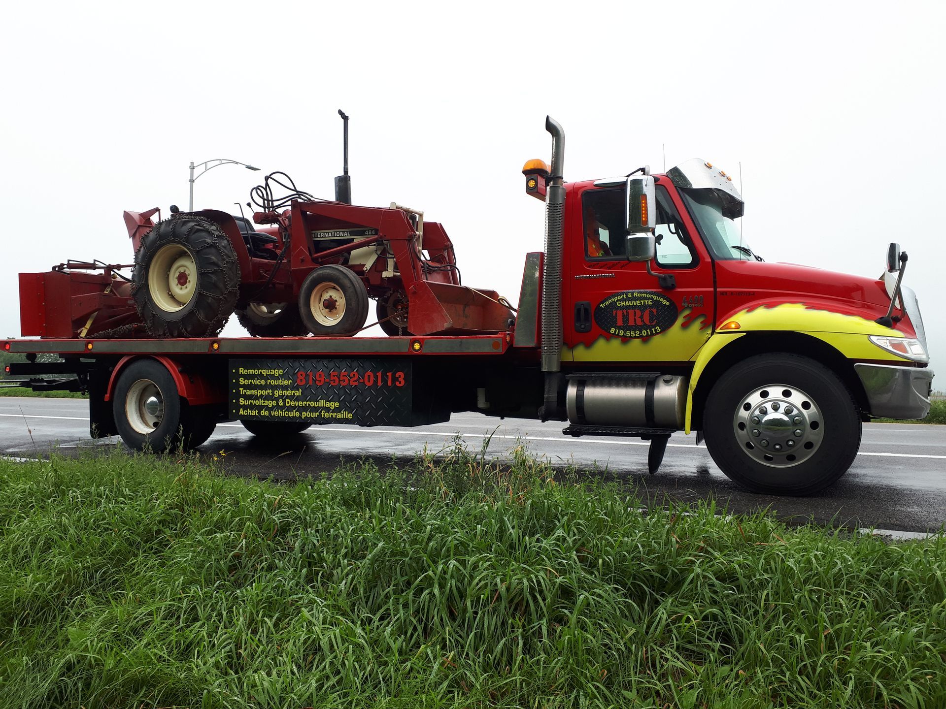 Une dépanneuse rouge transporte un tracteur rouge sur une remorque à plateau, au bord d'une route mouillée bordée d'herbe.