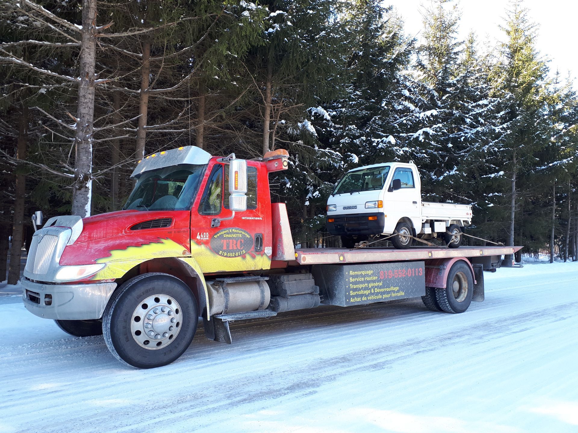 Une dépanneuse rouge transporte un petit camion blanc sur une route enneigée près d'une forêt.