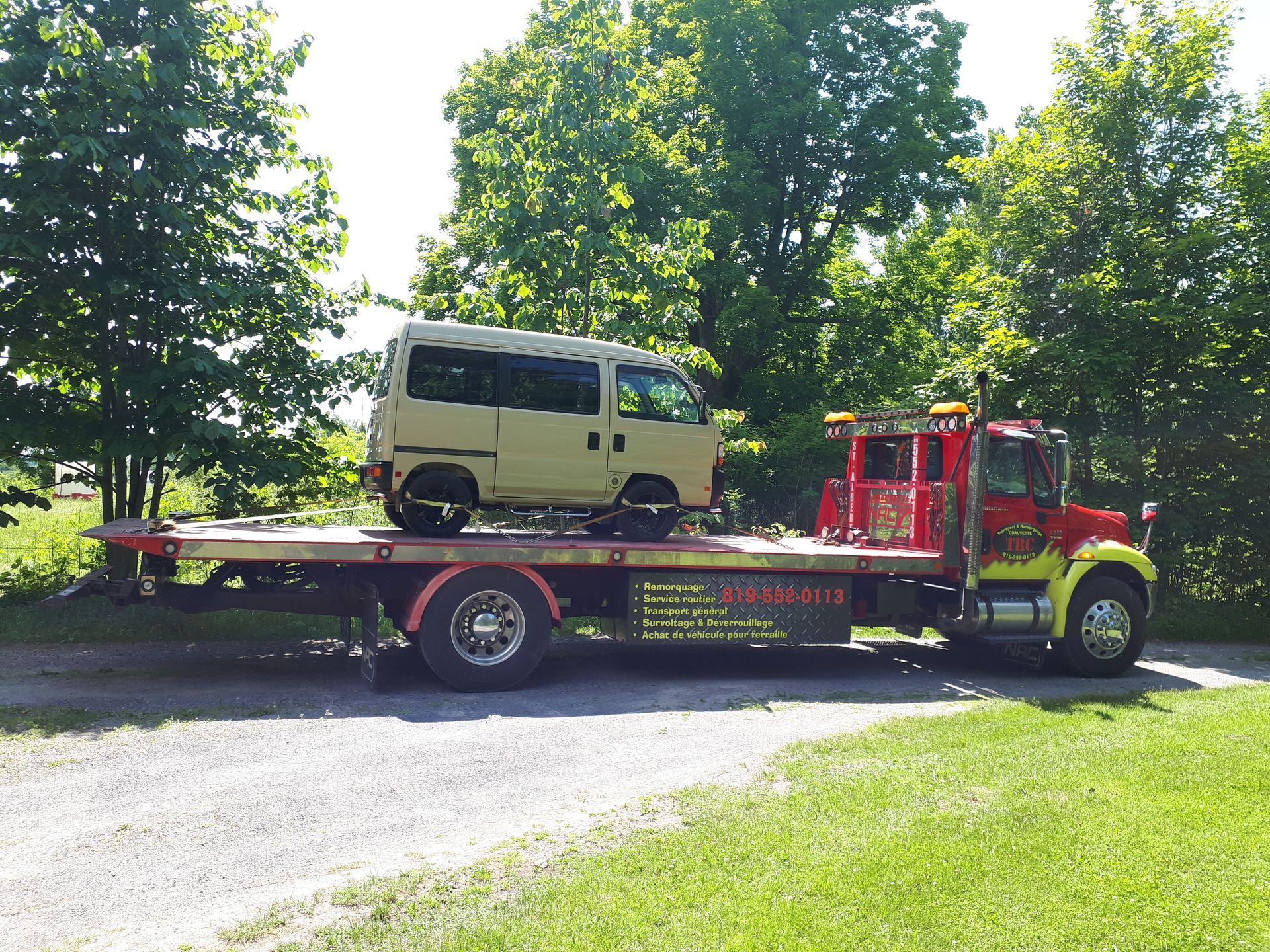 Une minifourgonnette beige chargée à l'arrière d'une dépanneuse rouge et jaune, garée sur une allée de gravier.