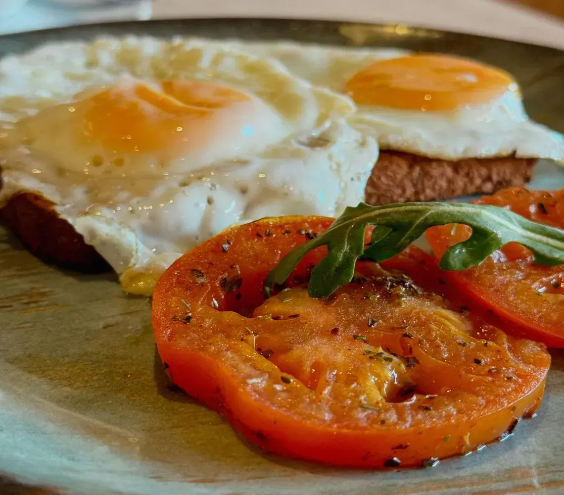 Two fried eggs on toast with seasoned tomato slices and a sprig of arugula on a ceramic plate.