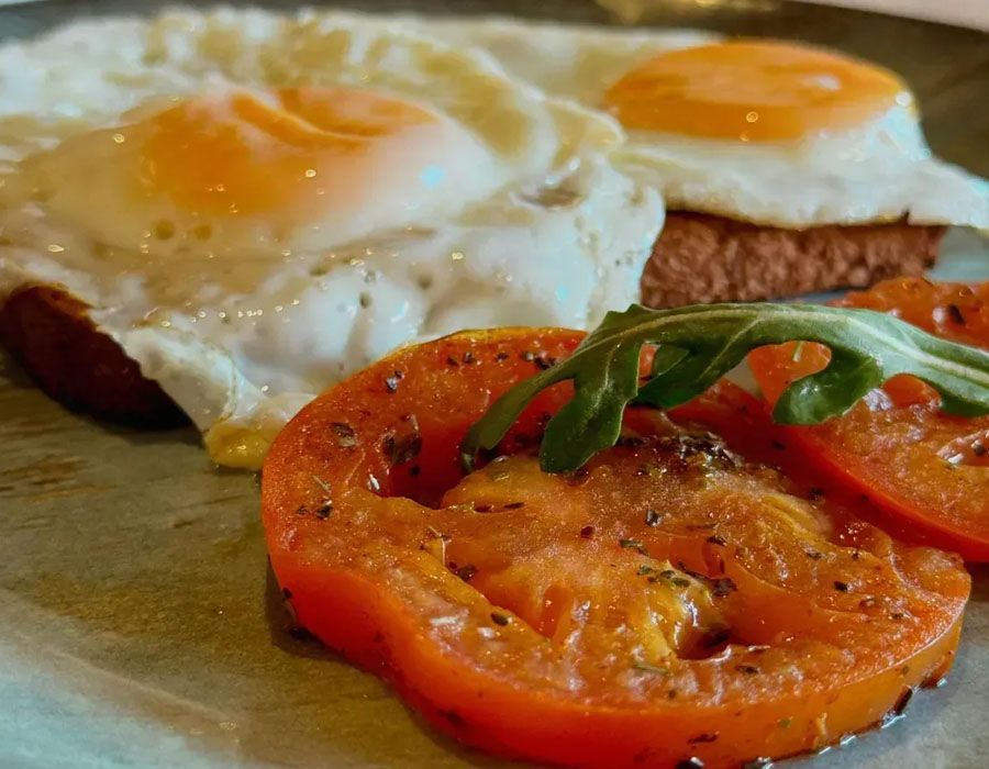 Two fried eggs on toast with seasoned tomato slices and a sprig of arugula on a ceramic plate.