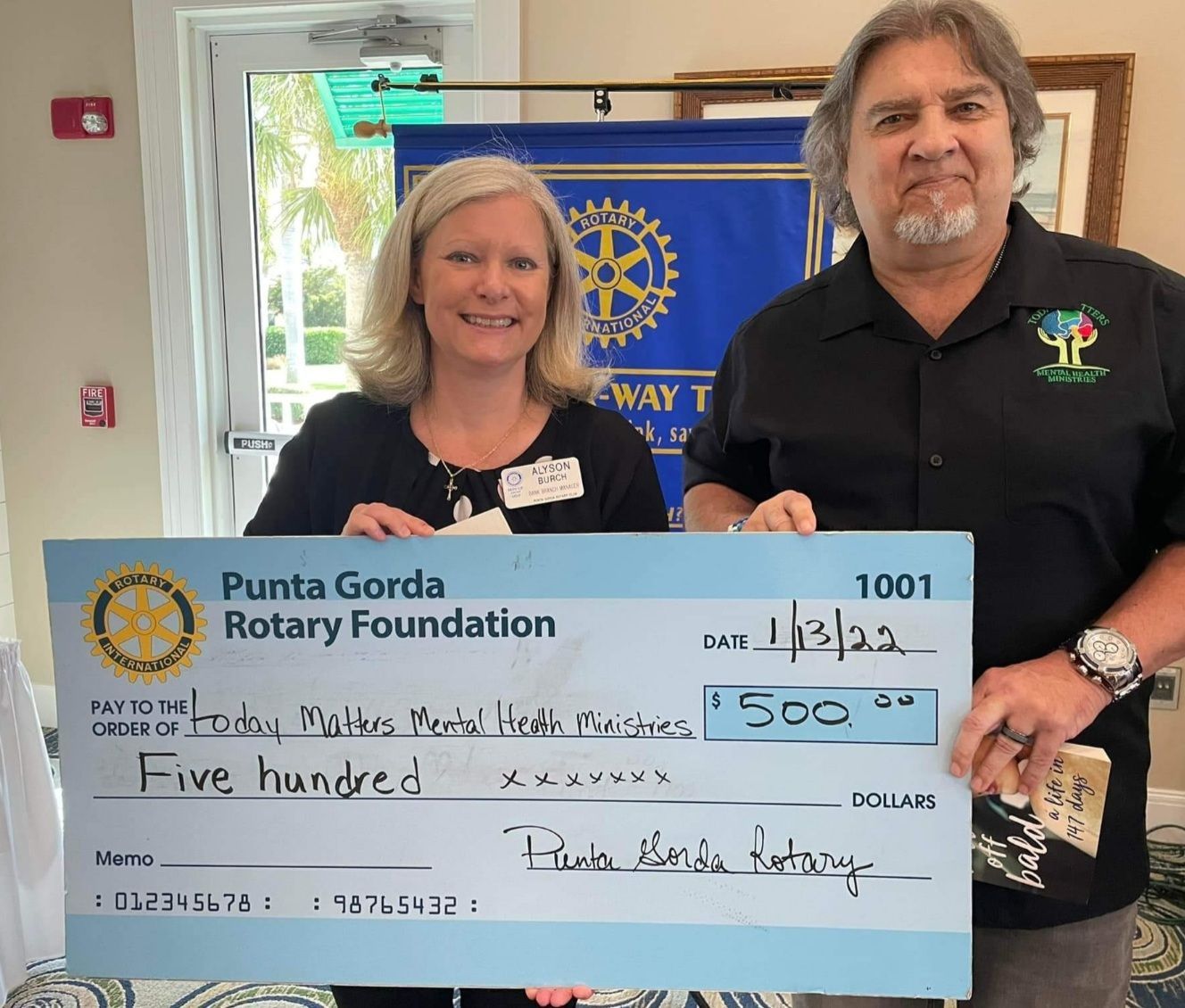 Two people hold a large Punta Gorda Rotary Foundation check for $500 in front of a Rotary banner.