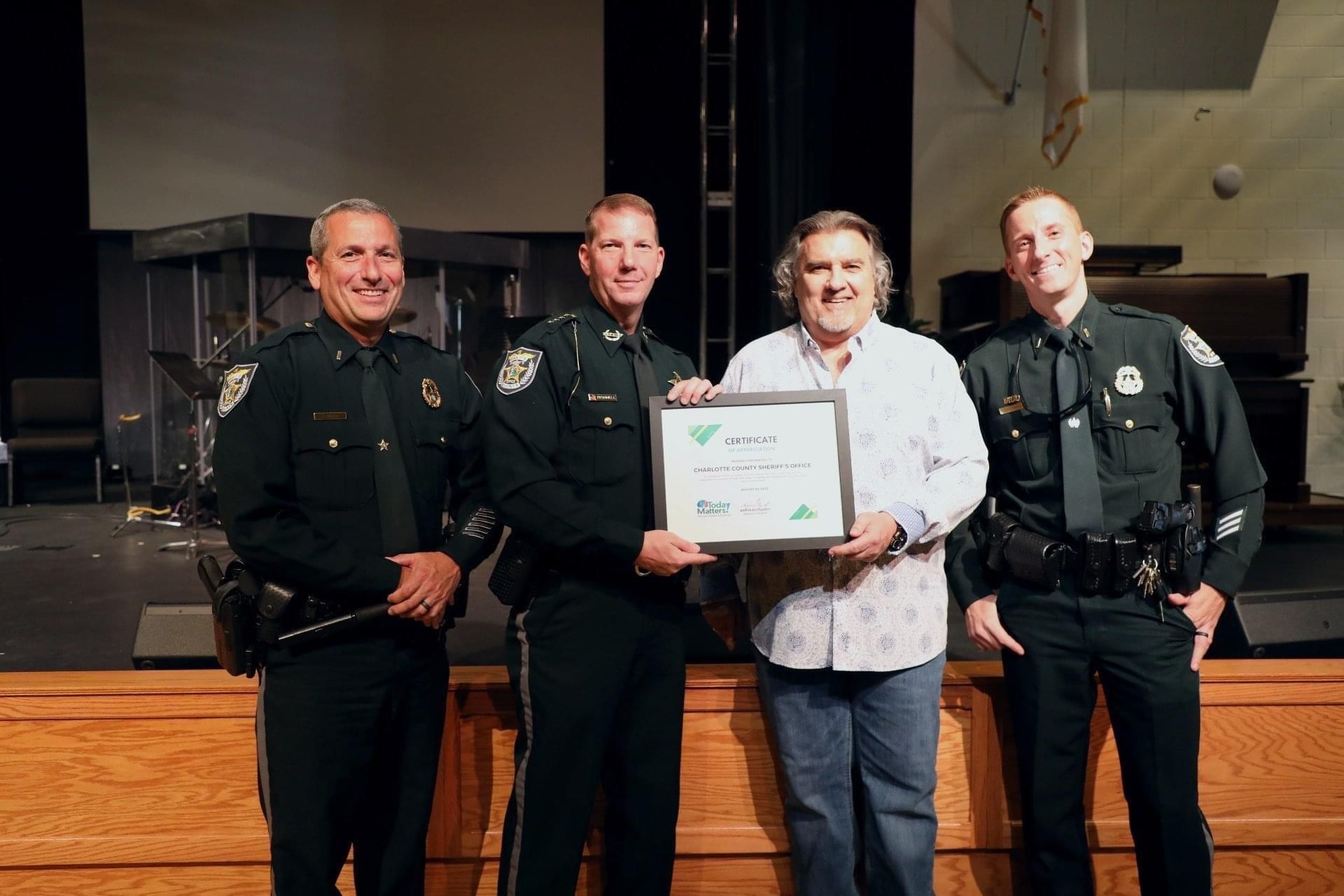 Four people on a stage, two uniformed officers flanking a woman holding a certificate, all smiling.