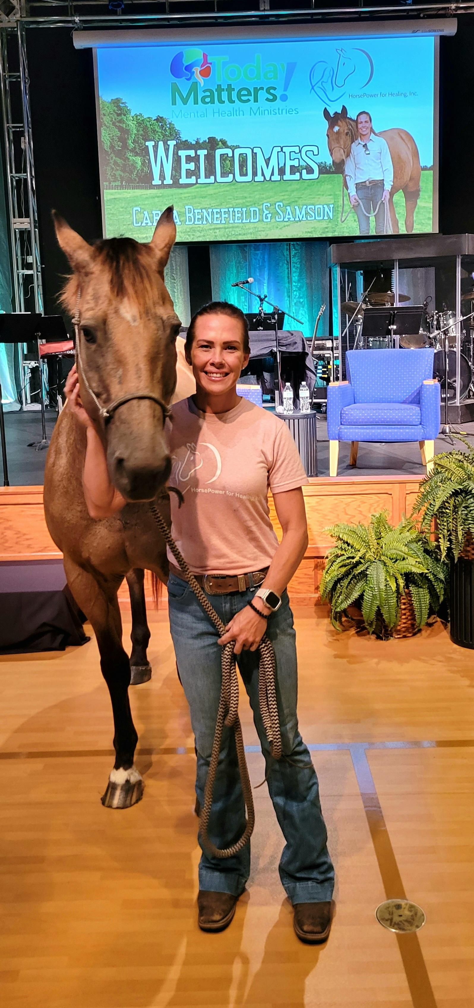 Man standing beside a horse on a stage with a blue “Welcome” sign and plants in the background