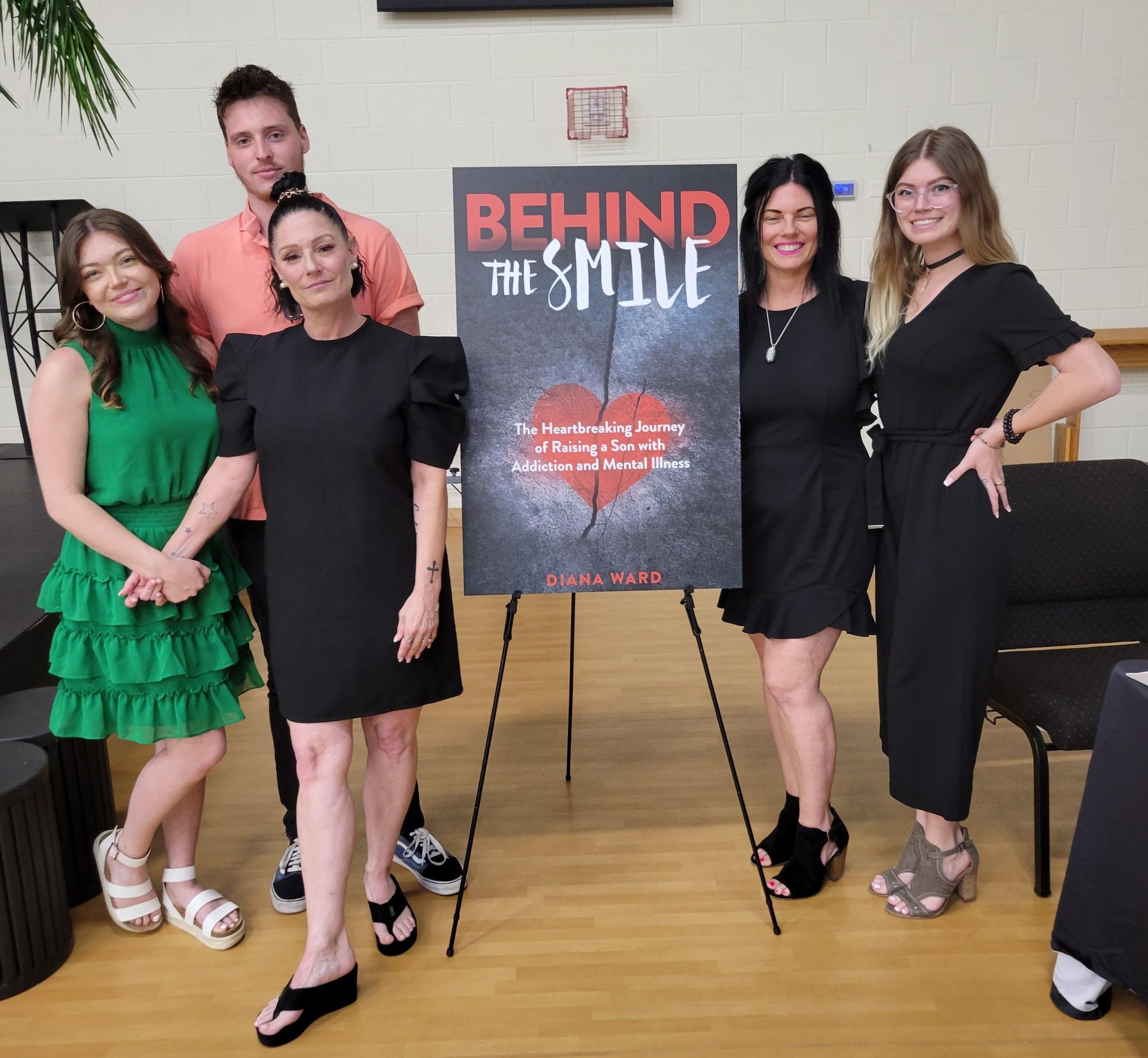 Five people posing beside a “Behind the Style” poster on easels in a room with wood floors.