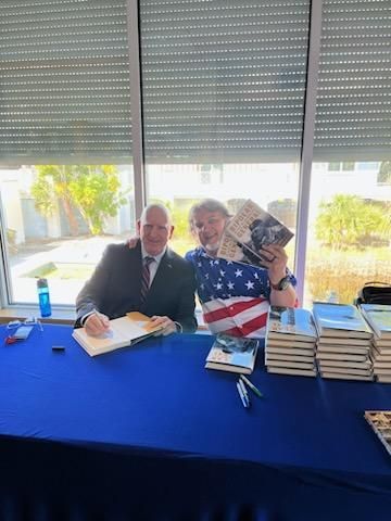 Two people at a blue table with books and an American flag backdrop, smiling and posing with signed books