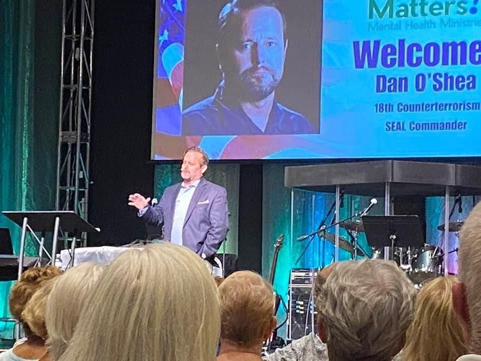 Speaker on stage at conference, with “Welcome Dan O’Shea” displayed on large screen behind him.