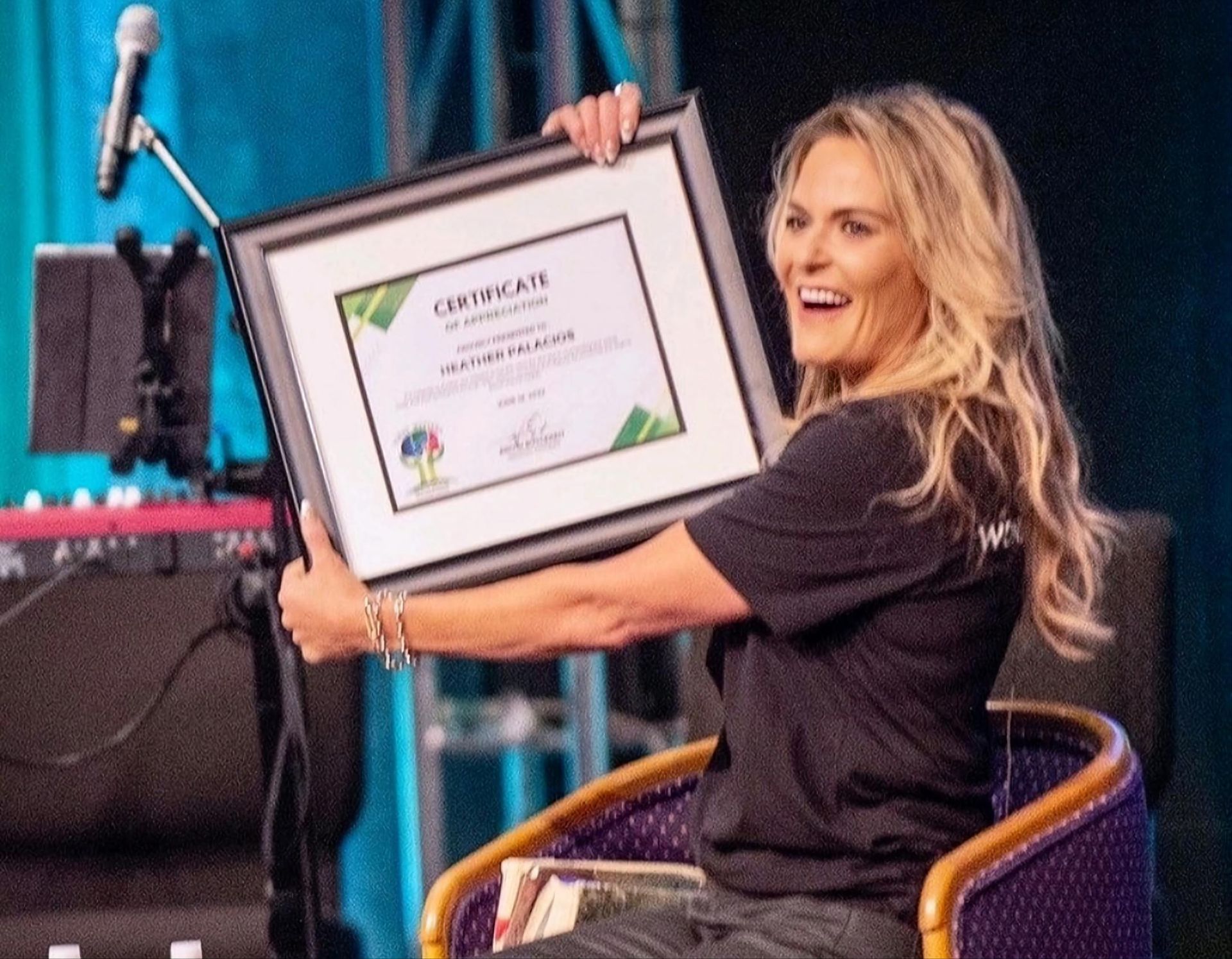 Woman holding a framed certificate on stage, smiling beside a microphone and chair