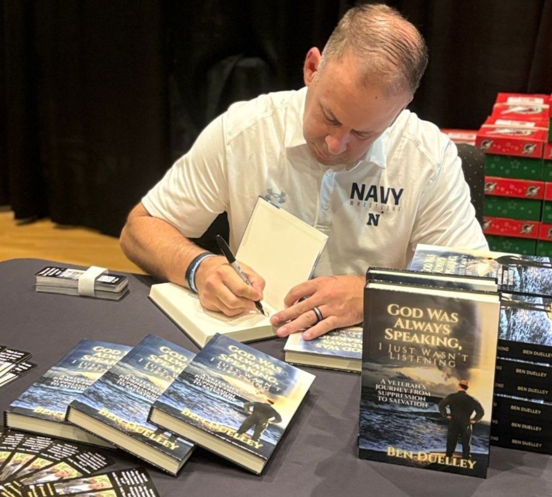 Man in white Navy shirt signing books at a table with stacks of books and a display copy nearby