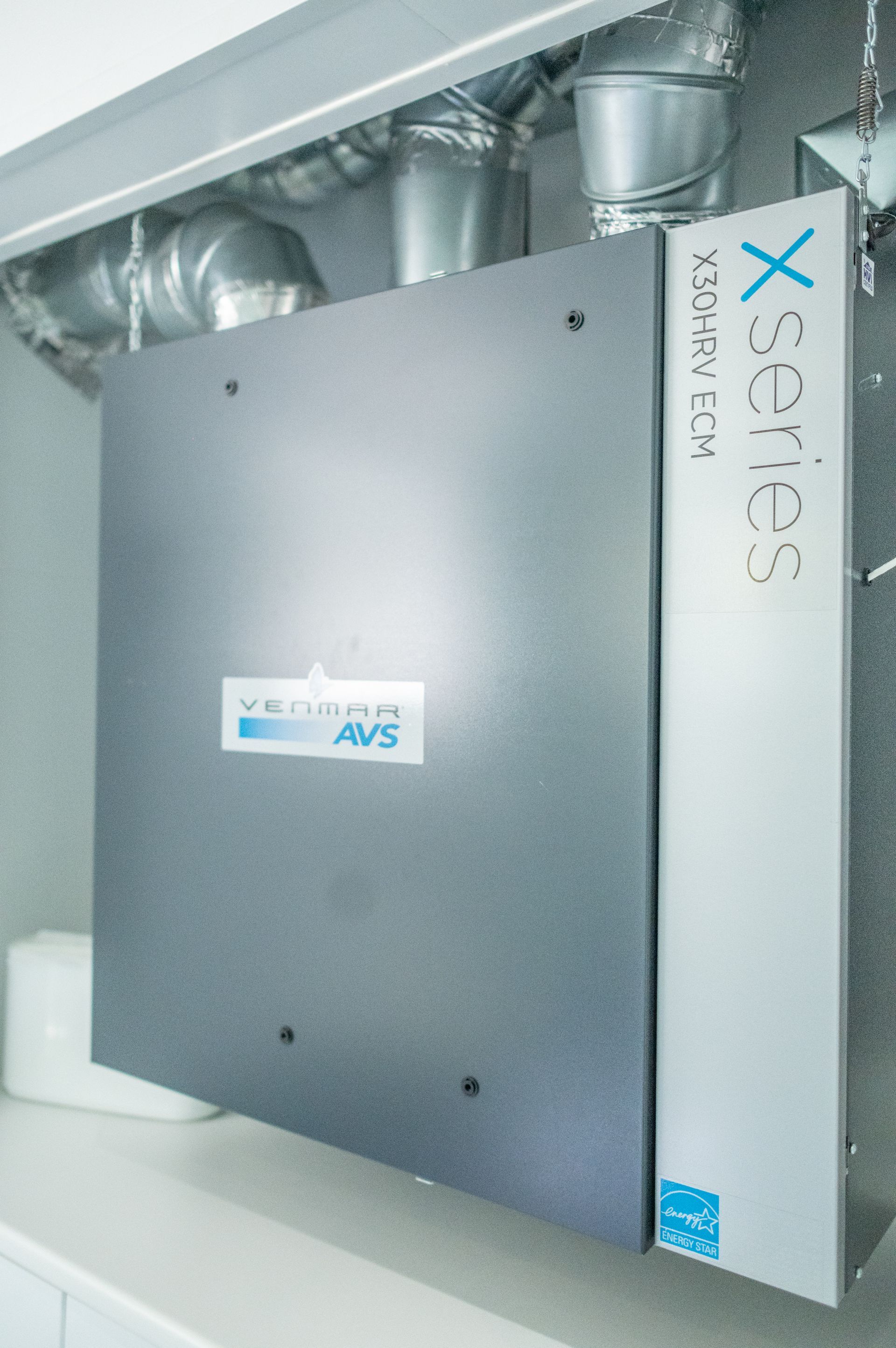 Gray air purifier, mounted on a wall. It has ductwork attached. The brand is written on the side.
