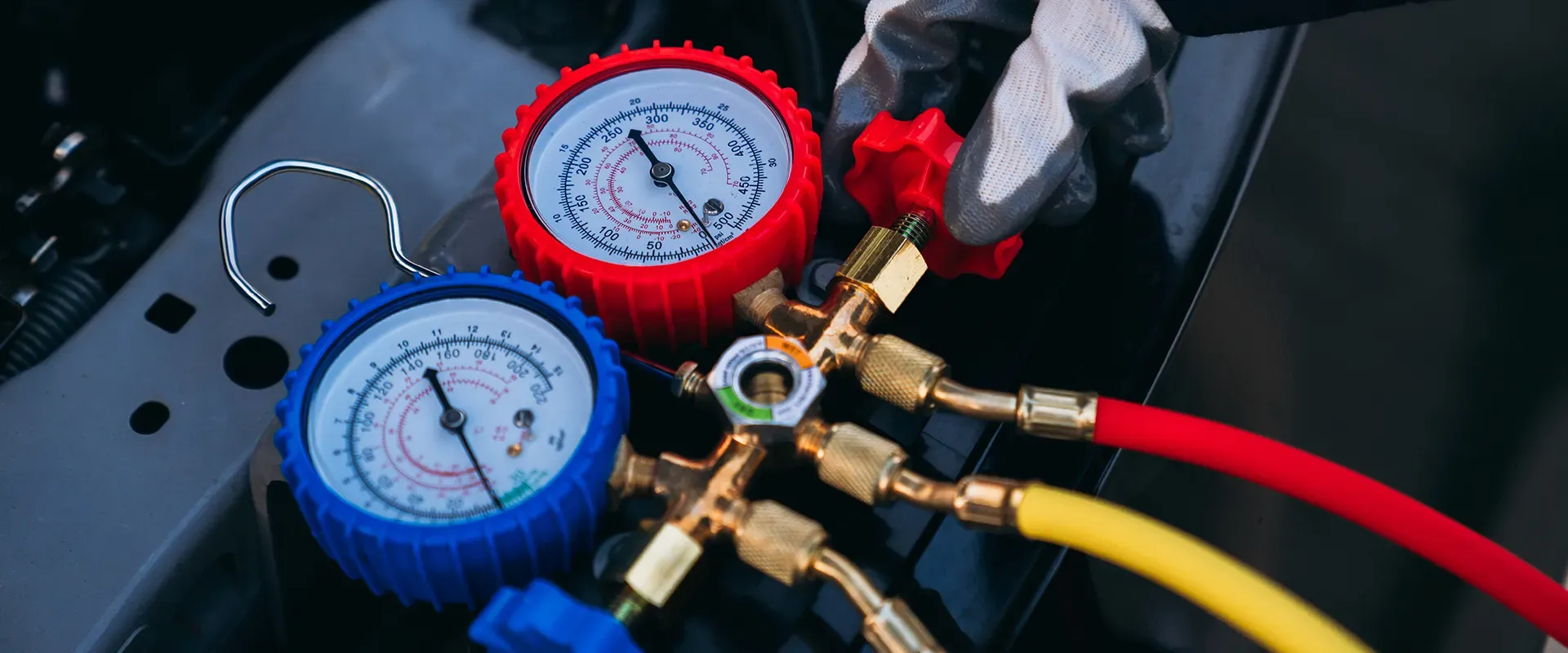 Close-up of gauges and hoses connected to a vehicle's AC system, being handled by a gloved hand.