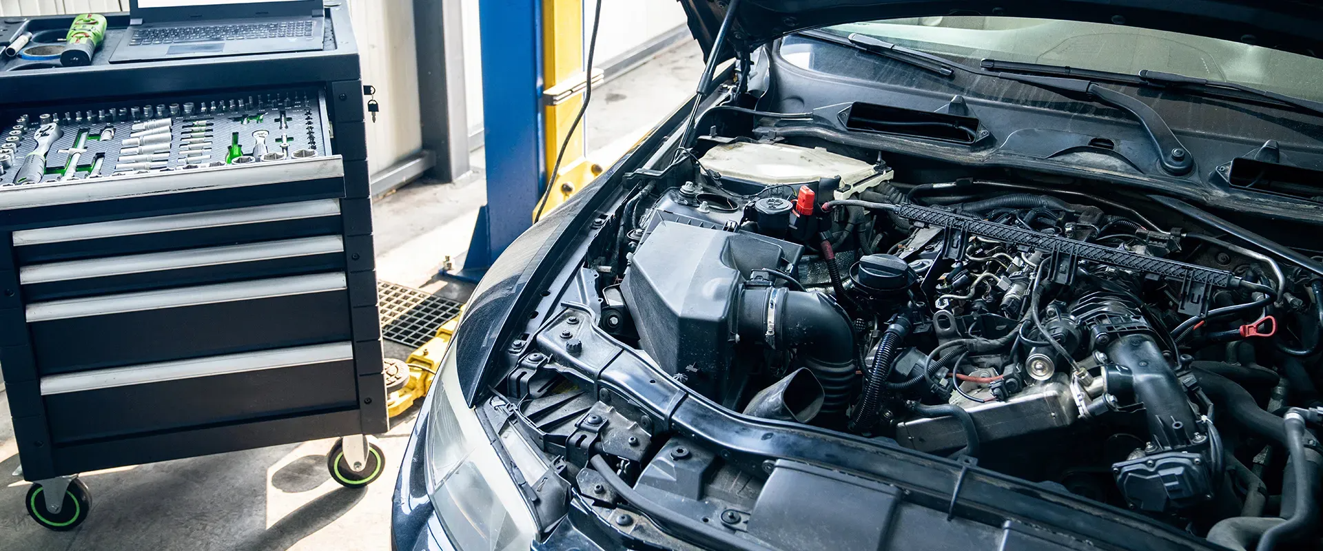 Car engine with open hood in a garage, next to a toolbox with tools.